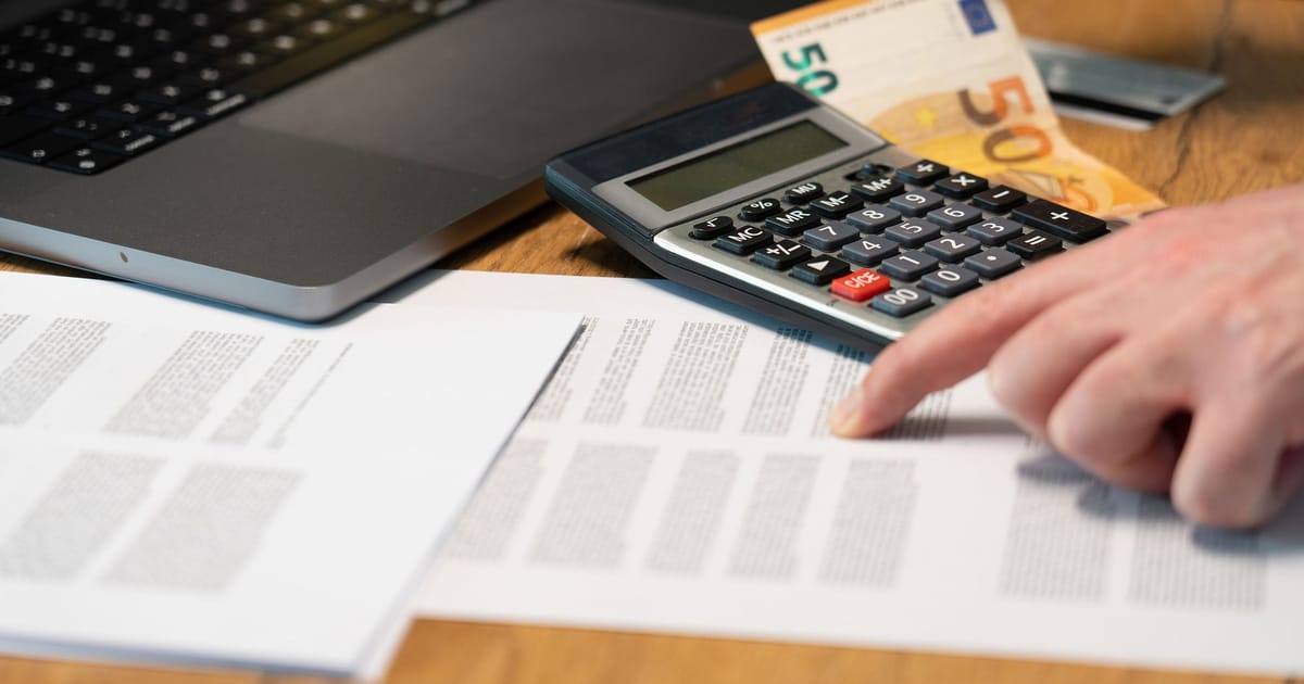 A hand working a calculator beside euro notes, a laptop and printed figures.