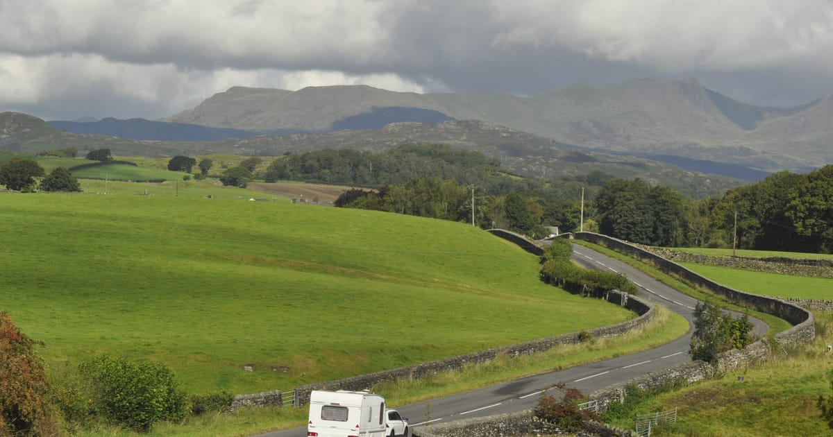 A car towing a caravan on a country road below hills.